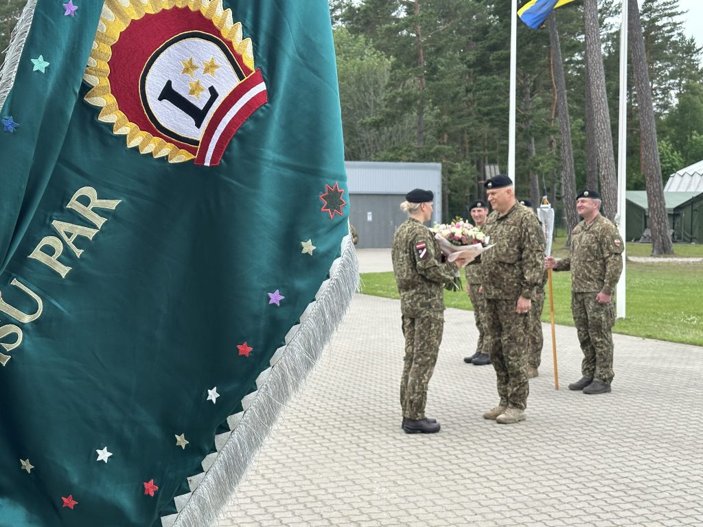 Vīrietis militārā formā pasniedz ziedus sievietei militārā formā, fonā redzams zaļš karogs.
