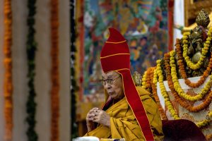 Tibetas trimdas garīgais līderis Dalailama sēž, tērpies safrāna dzeltenā apmetnī un sarkanā cepurē, ar brilles un sakļautām rokām