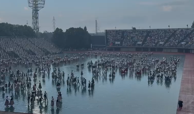 Liels dejotāju pulks stadionā, kas klāts ar ūdeni no lietus, tribīnes ar skatītājiem fonā.