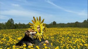 Cilvēks ar pienenei līdzīgu masku un suns pļavā pilnā ar pienenēm