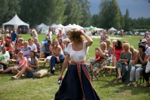 Sieviete tautiskā tērpā ar vainagu soļo publikas priekšā brīvdabas festivālā