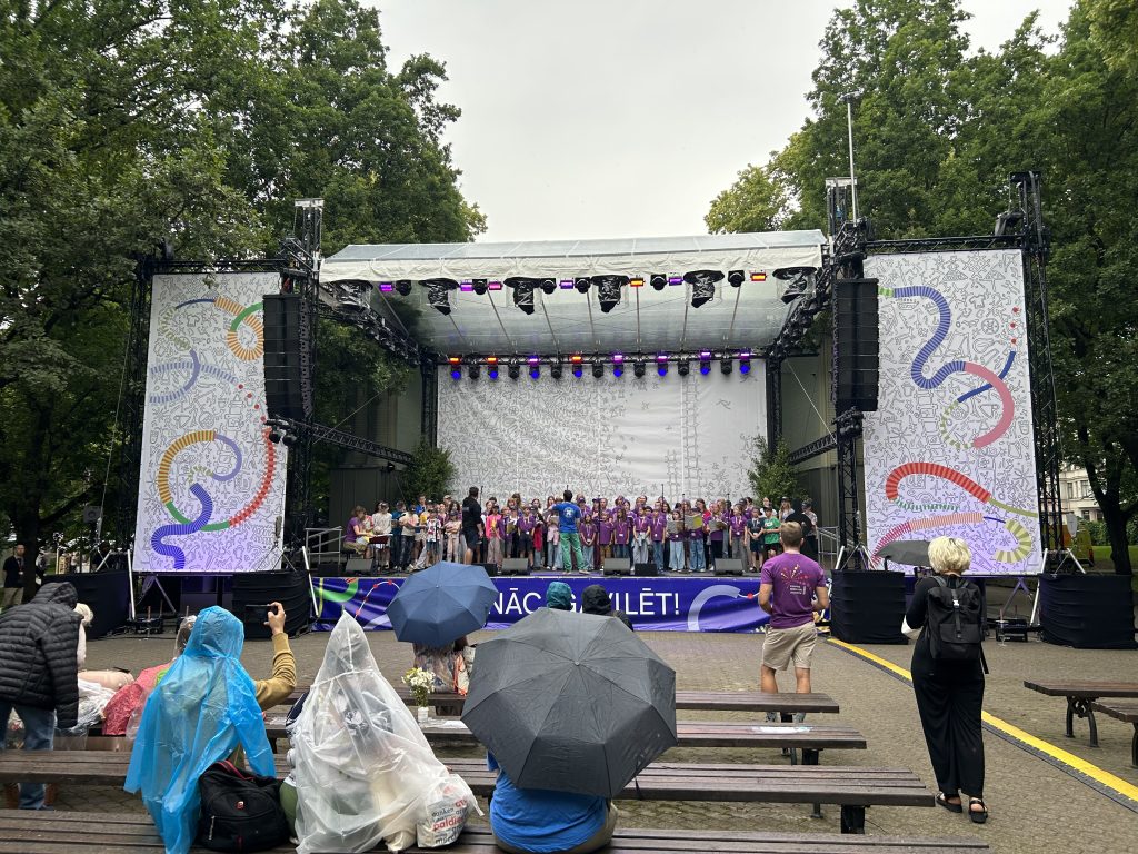 Bērnu koris violetos kreklos dzied uz skatuves Vērmanes dārzā