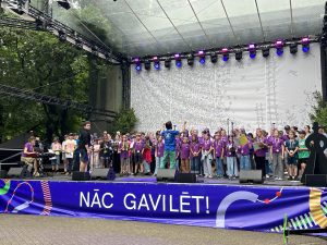 Liels bērnu koris violetos T-kreklos uzstājas uz skatuves Vērmanes dārzā, diriģents paceļ rokas.