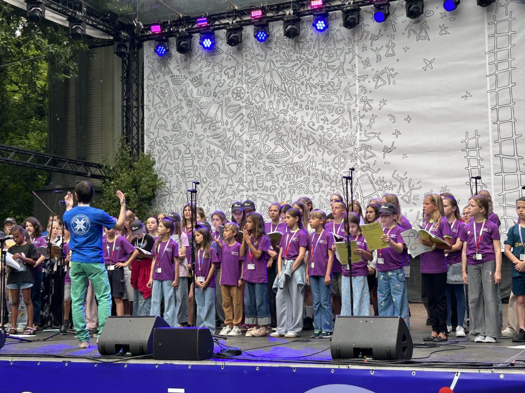 Bērnu koris violetos kreklos dzied uz skatuves, diriģents ar muguru pret skatītājiem.