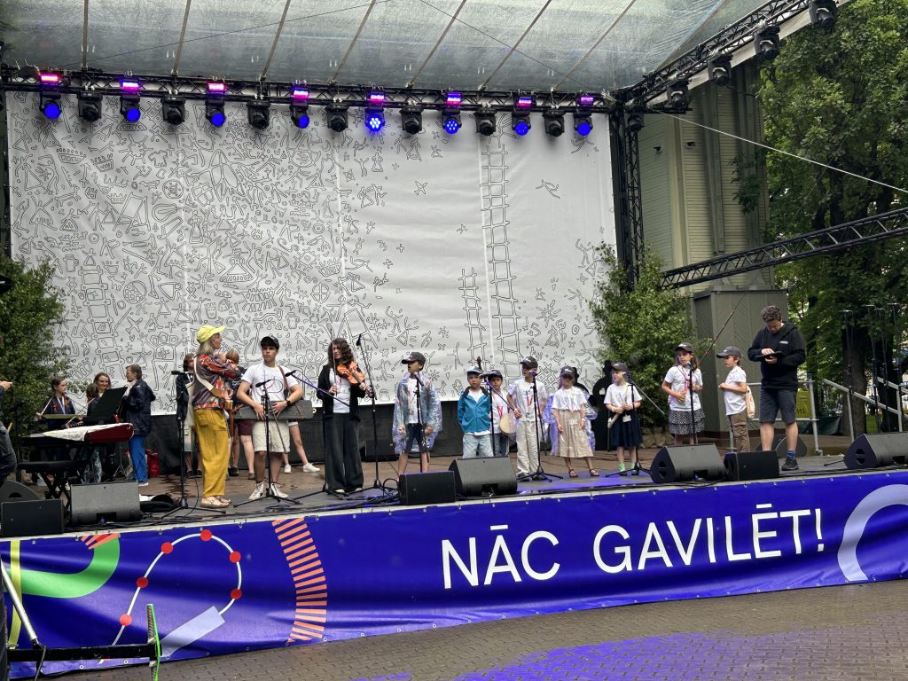 Skolotāja dzeltenā cepurē un jauniešu grupa ar mūzikas instrumentiem uzstājas uz skatuves.