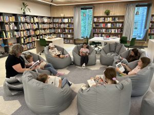 Bērni un pieaugušais sēž aplī pelēkos sēžammaisos un kopīgi lasa bibliotēkā