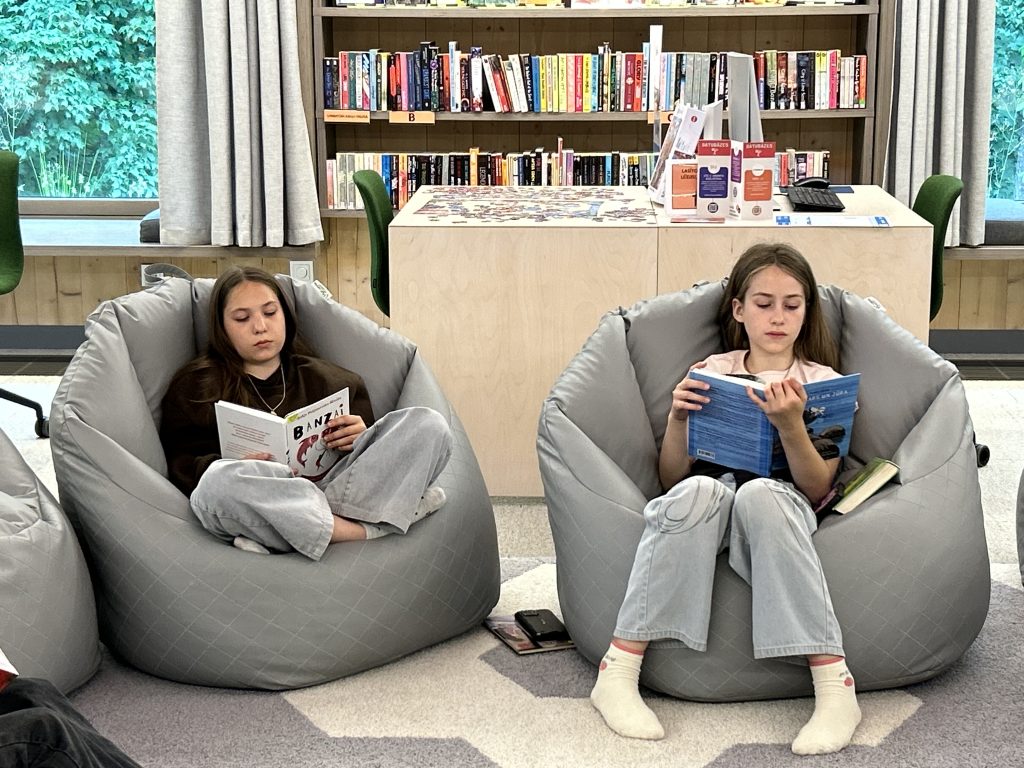 Divas meitenes ērti iekārtojušās sēžammaisos un lasa grāmatas bibliotēkā.