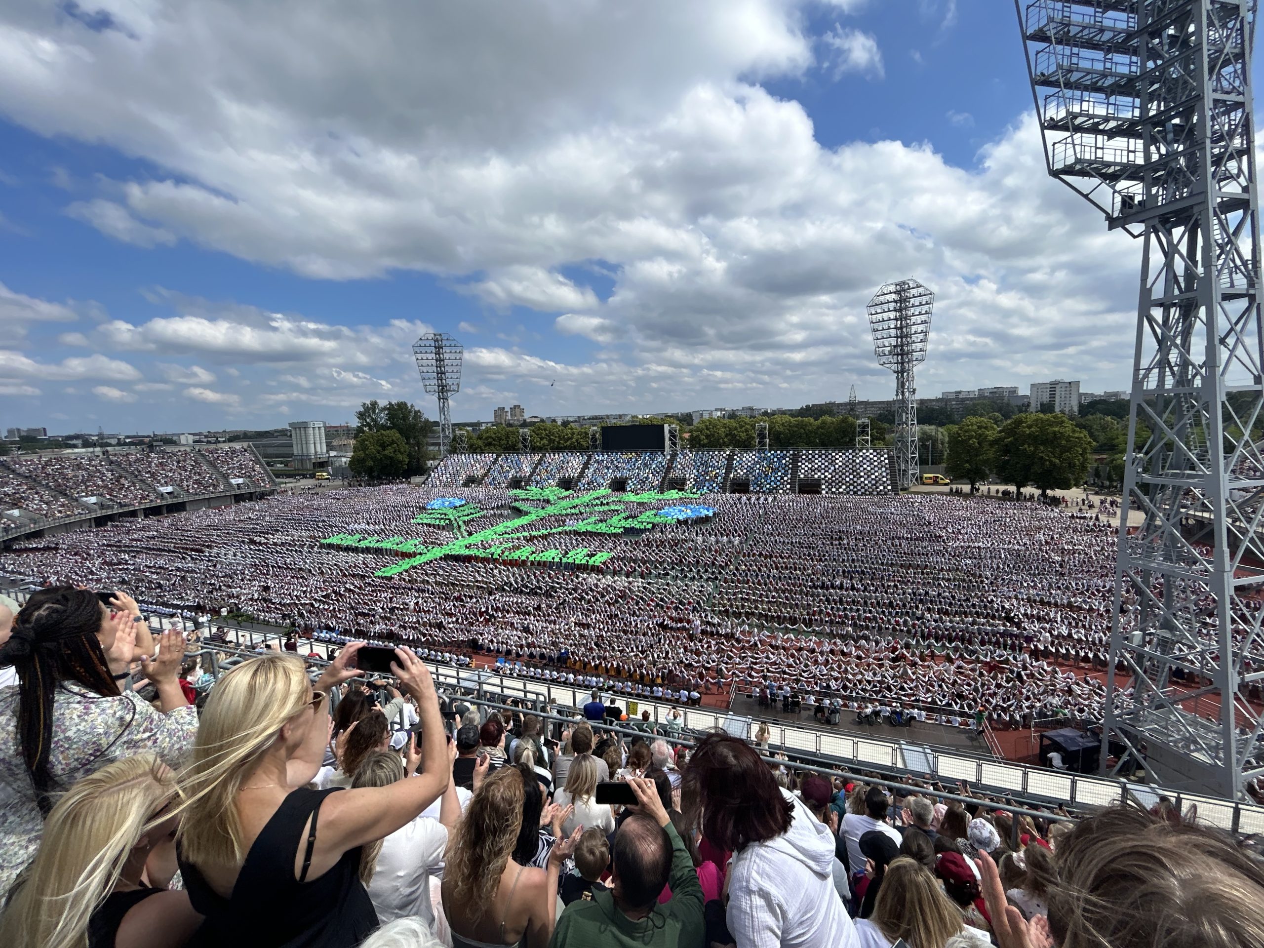 Daugavas stadions piepildīts ar dejotājiem tradicionālos tērpos, kas veido lielu, zaļu tautas deju rakstu.
