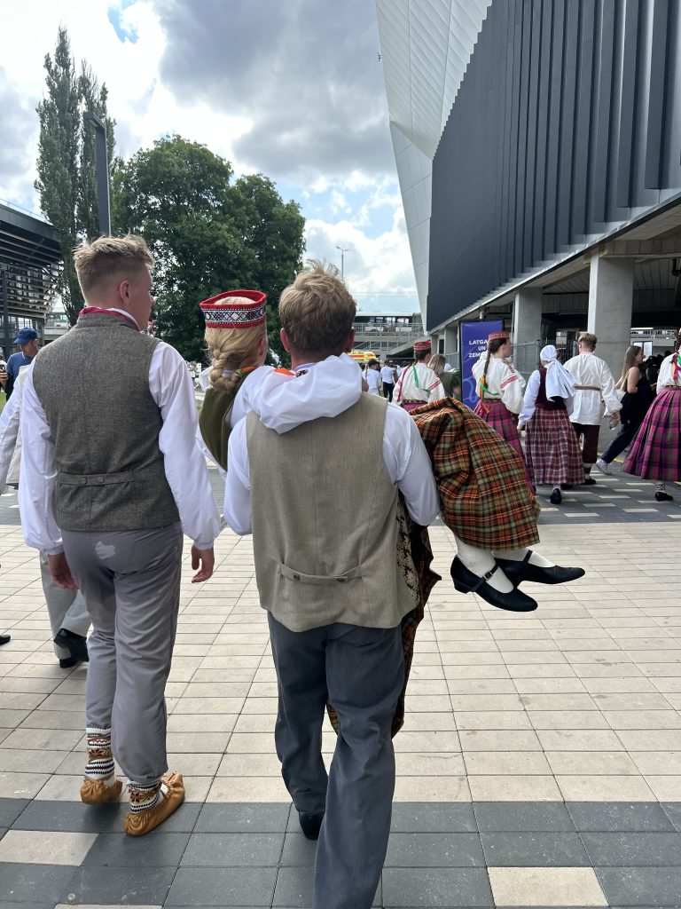 Jauniešu dejotāji tautas tērpos iet gar modernu ēku, viens jaunietis nes meiteni rokās.