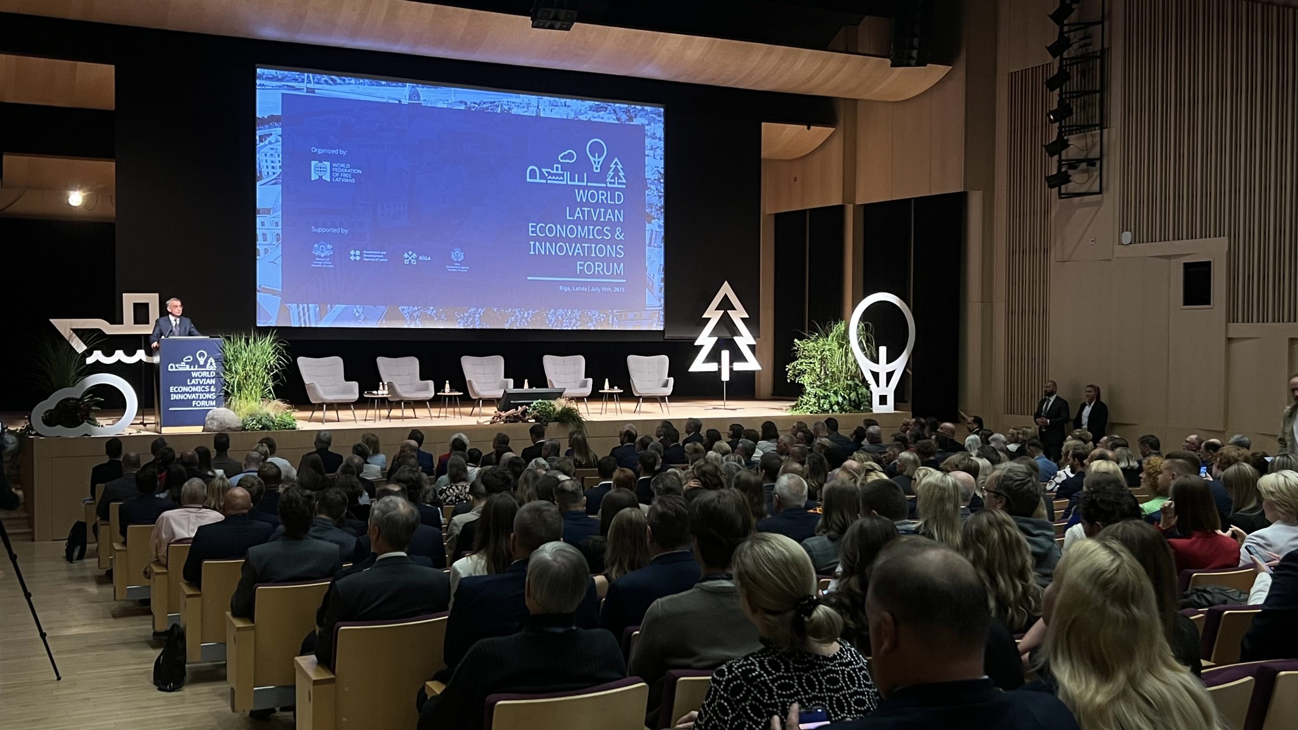 Plašs skats uz Pasaules latviešu ekonomikas un inovāciju foruma zāli ar runātāju uz skatuves un lielu auditoriju