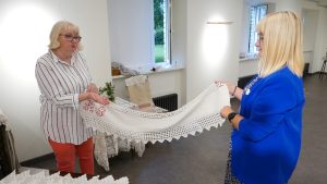 Divas sievietes tur izšūtu dvieli ar mežģīnēm izstādē