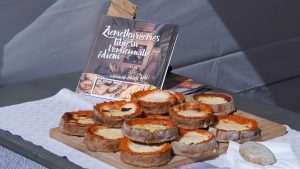 Uz koka paplātes izkārtoti svaigi cepti, zeltaini sklandrauši ar burkānu un kartupeļu pildījumu, fonā redzama grāmata "Ziemeļkurzemes lībiešu tradicionālie ēdieni"