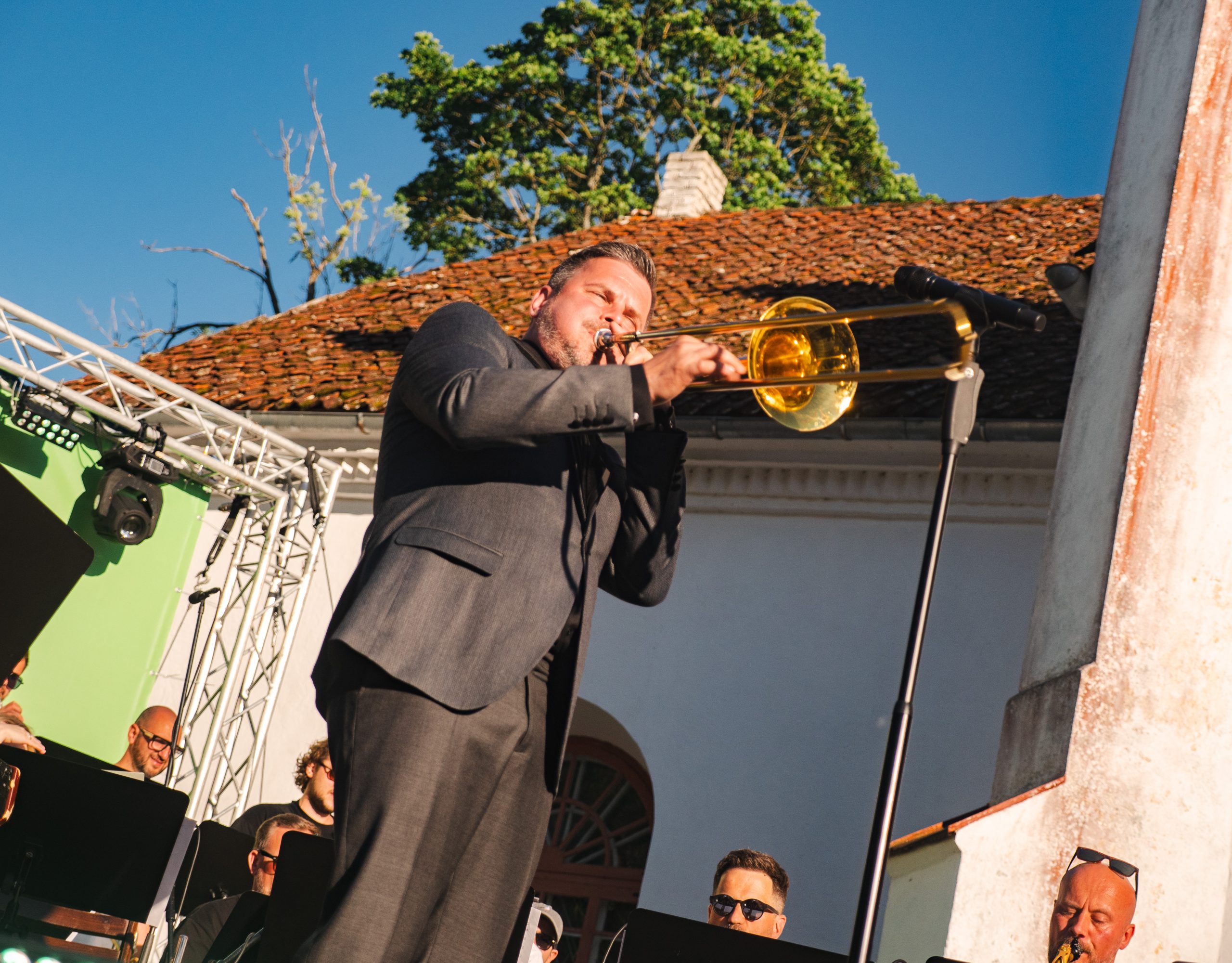 Intars Busulis spēlē trombonu uz skatuves Talsos saulainā dienā.