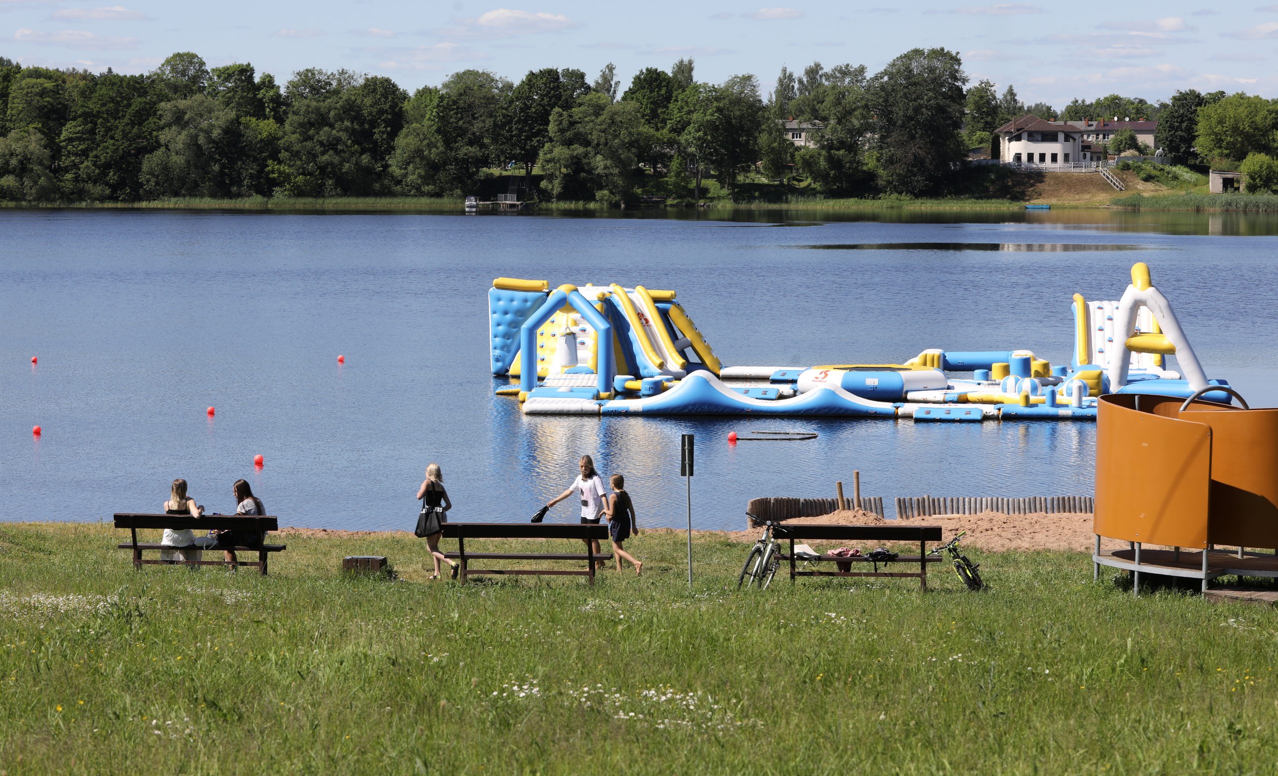 Ūdens atrakciju parks ezerā un cilvēki uz soliņiem pļavā pie krasta.