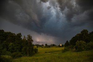 Tumši negaisa mākoņi un zibens virs Latvijas lauku ainavas.