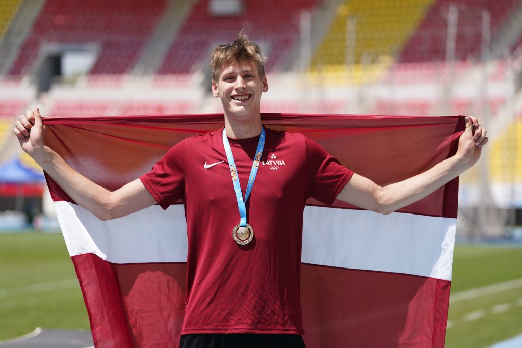 Jauns vīrietis sarkanā sporta kreklā ar medaļu ap kaklu un Latvijas karogu, smaidot stāv sporta stadionā.
