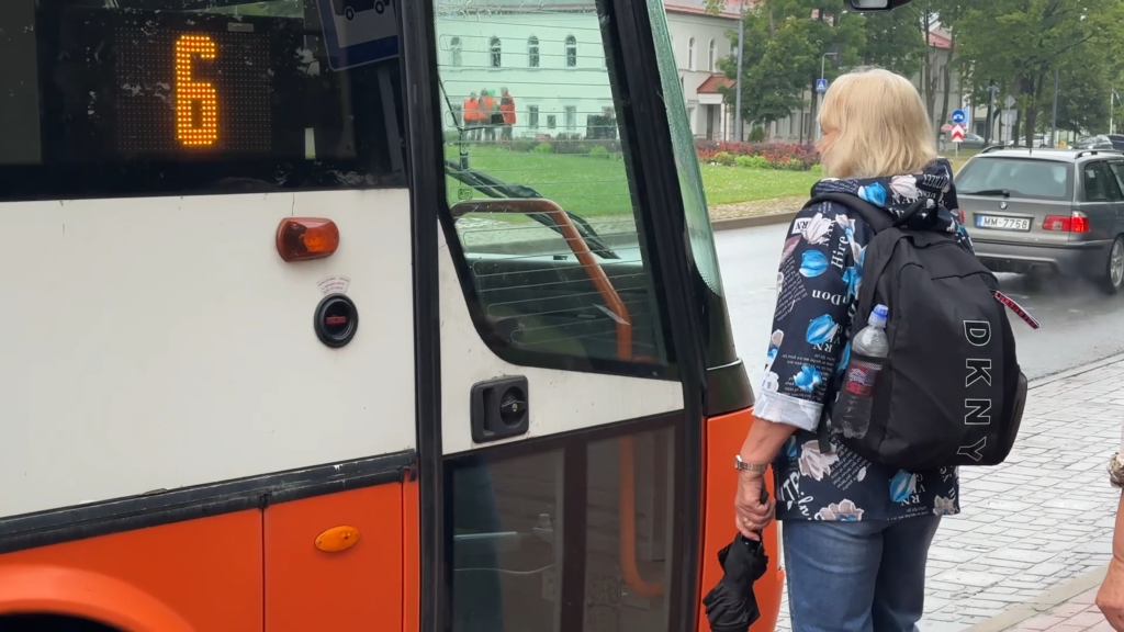 Sieviete ar melnu DKNY mugursomu un lietussargu stāv pie oranža autobusa durvīm.