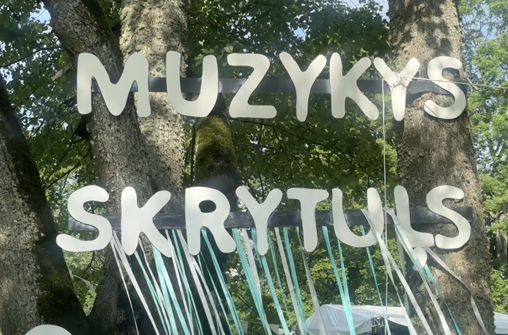 Uz koka piestiprinātas lielas baltas burtu zīmes "MUZYKYS SKRYTUĻS"