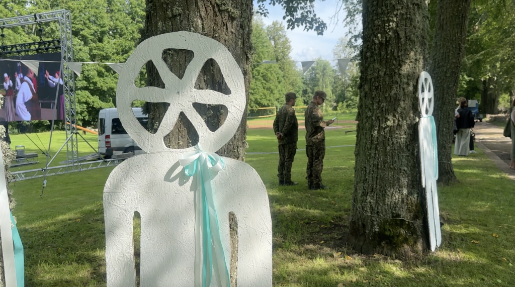 Baltas figūras ar ratiem galvas vietā un lentām pie koku stumbriem