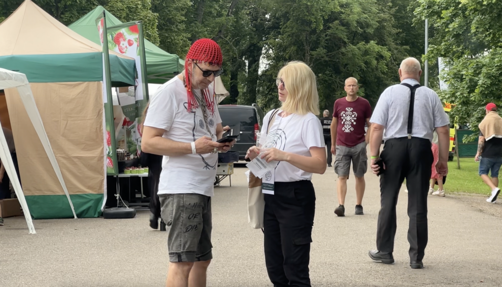 : Vīrietis sarkanā cepurē un sieviete stāv un sarunājas festivāla zonā