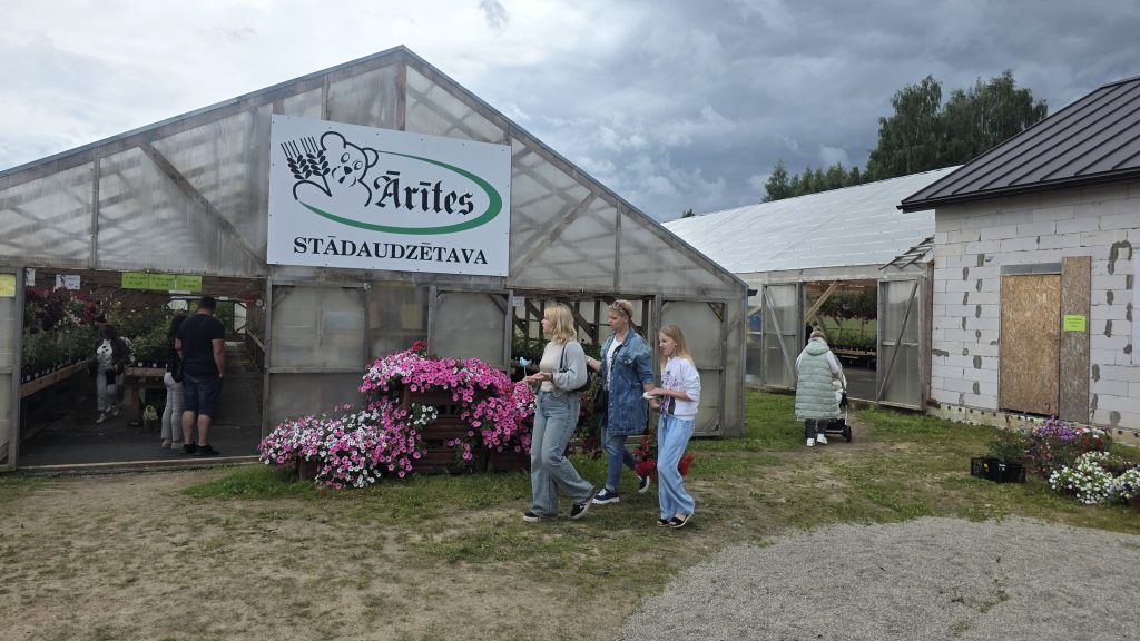 Trīs sievietes stāv pie siltumnīcas ieejas, ko ieskauj ziedošas puķes