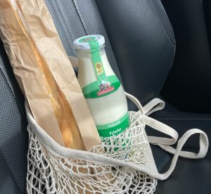 Stikla piena pudele un bagete režģveida maisiņā uz automašīnas sēdekļa.