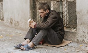 Cilvēks sēž uz kartona gabala pie ēkas sienas, rokās turot papīra glāzi, kas liecina par lūgšanu pēc palīdzības.