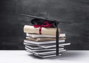 Diploms ar sarkanu lenti un akadēmiskā cepure novietoti uz grāmatu kaudzes melnas tāfeles priekšā
