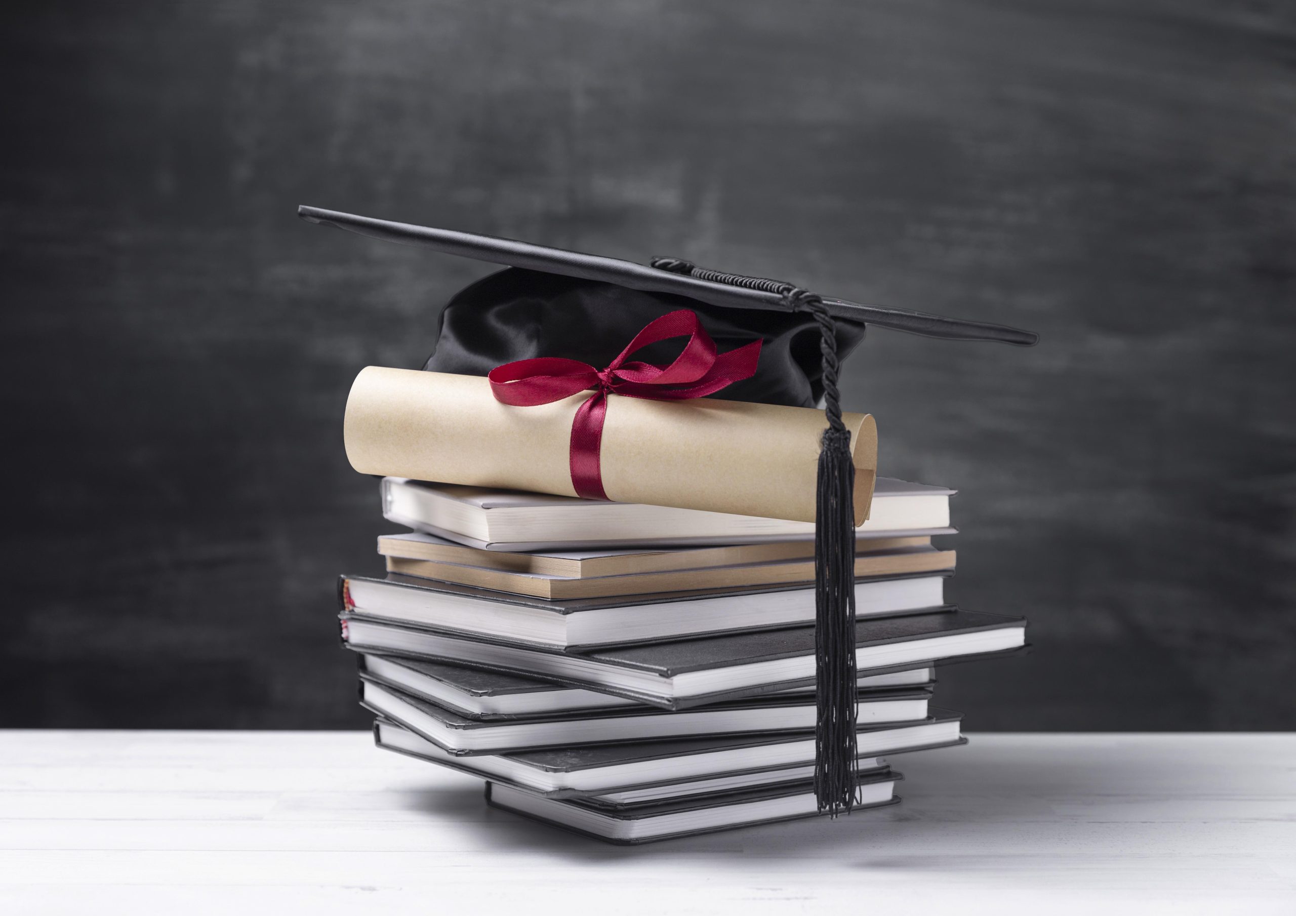 Diploms ar sarkanu lenti un akadēmiskā cepure novietoti uz grāmatu kaudzes melnas tāfeles priekšā