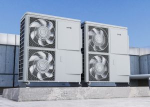 Divas pelēkas āra gaisa-ūdens siltumsūkņu vienības ar ventilatoriem, kas uzstādītas uz ēkas jumta vai pamatnes zem skaidrām debesīm.