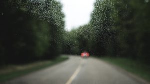 Lietus lāses uz automašīnas loga ar izplūdušu skatu uz ceļu un mežu