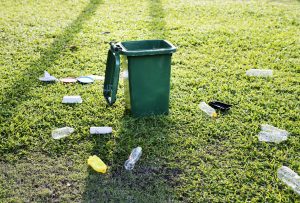 Zaļa atkritumu urna ar paceltu vāku, kas atrodas zaļā zālienā, ap kuru izmētāti dažādi plastmasas atkritumi un pudeles