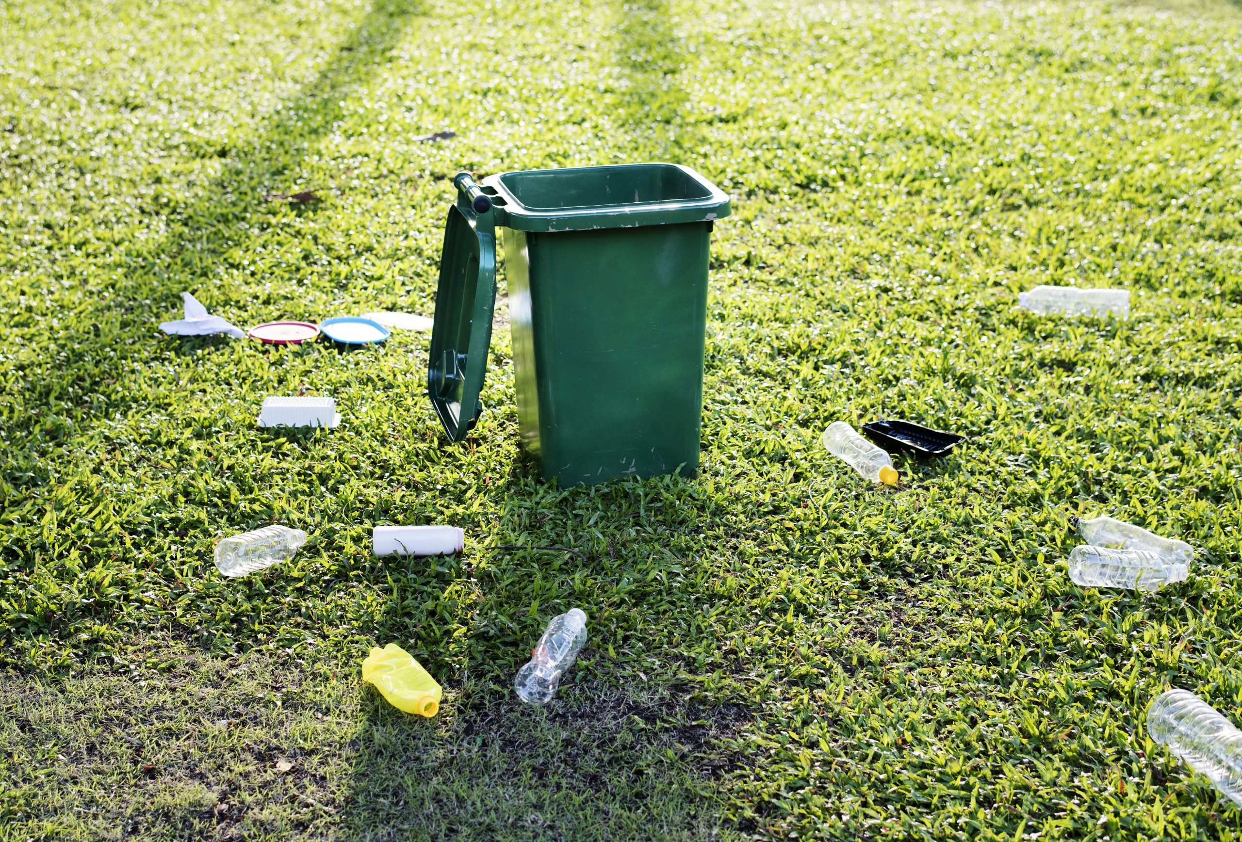 Zaļa atkritumu urna ar paceltu vāku, kas atrodas zaļā zālienā, ap kuru izmētāti dažādi plastmasas atkritumi un pudeles