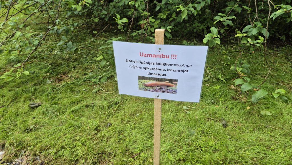Balts brīdinājuma plakāts uz koka staba ar uzrakstu par Spānijas kailgliemežu apkarošanu.