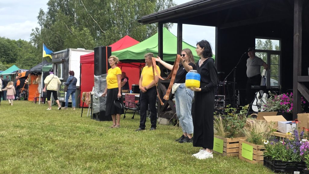 Vairāki cilvēki stāv uz zāles laukuma, fonā teltis un neliela skatuve. Divas sievietes tur Ukrainas karoga krāsas priekšmetus