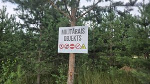 Brīdinājuma zīme ar uzrakstu "MILITĀRAIS OBJEKTS" un aizlieguma simboliem, kas piestiprināta pie koka mežā