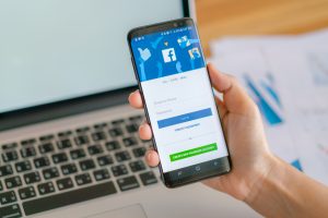 Roka tur Samsung Galaxy S8 viedtālruni ar atvērtu Facebook pieteikšanās ekrānu, fonā redzama klaviatūra un dokumenti.