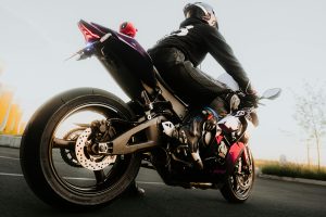 Motociklists pilnā ekipējumā sēž uz sporta motocikla, gatavs braucienam, ar zemu saules staru apgaismojumu.