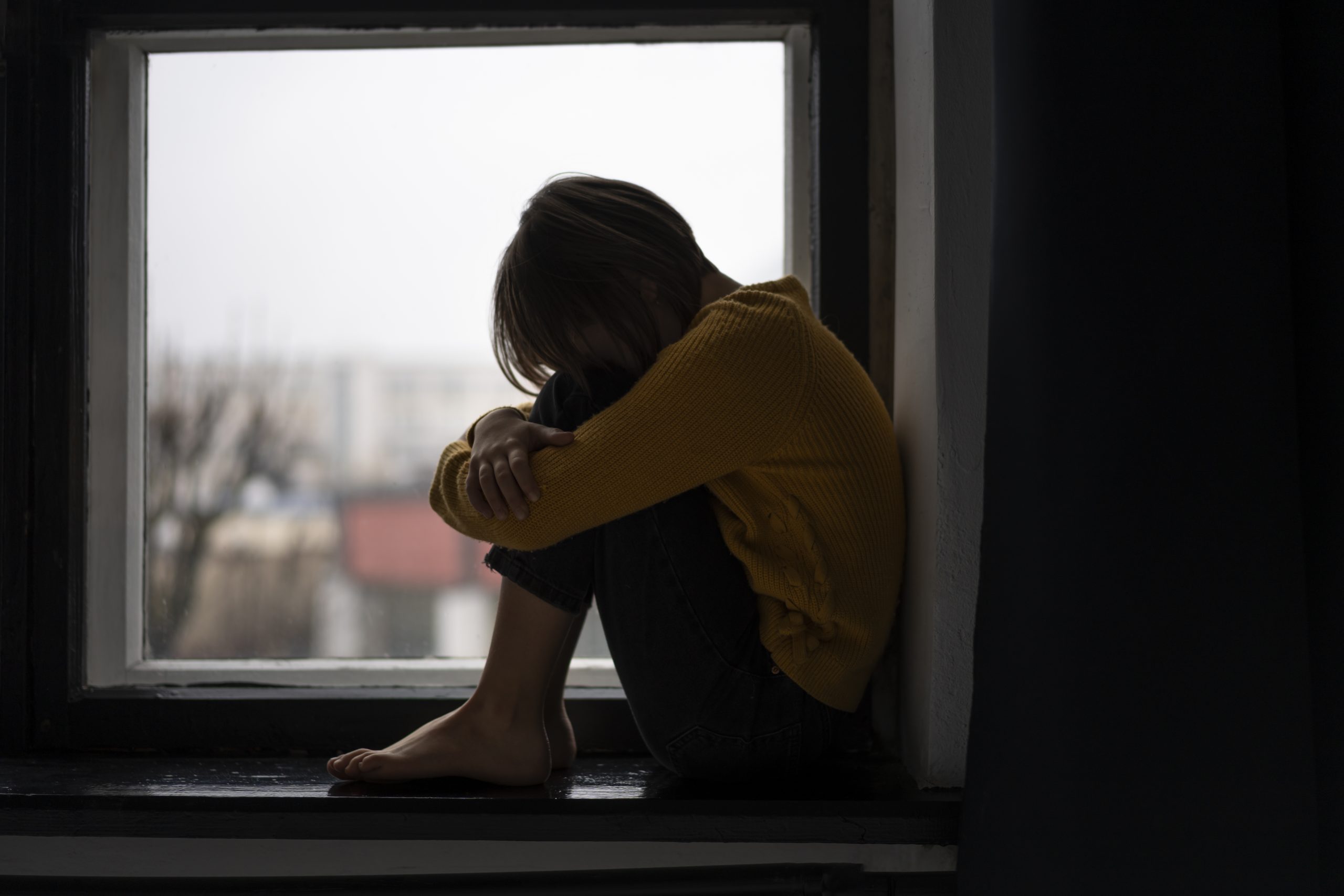 Skumja meitene sēž palodzes nišā, sakļāvusi rokas ap ceļiem.