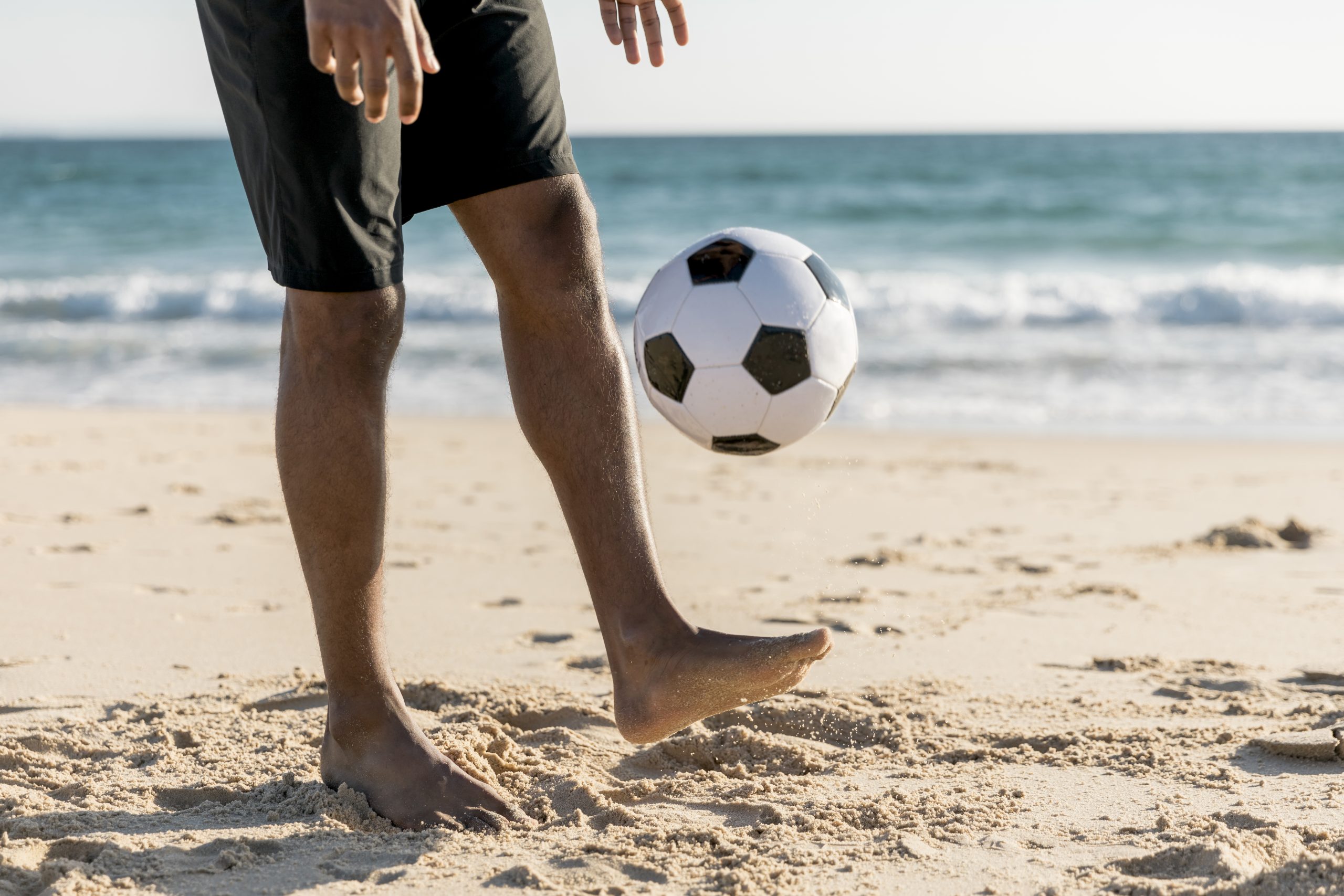 Persona pludmalē spēlējas ar futbola bumbu, veicot žonglēšanu ar kāju, ar jūru fonā