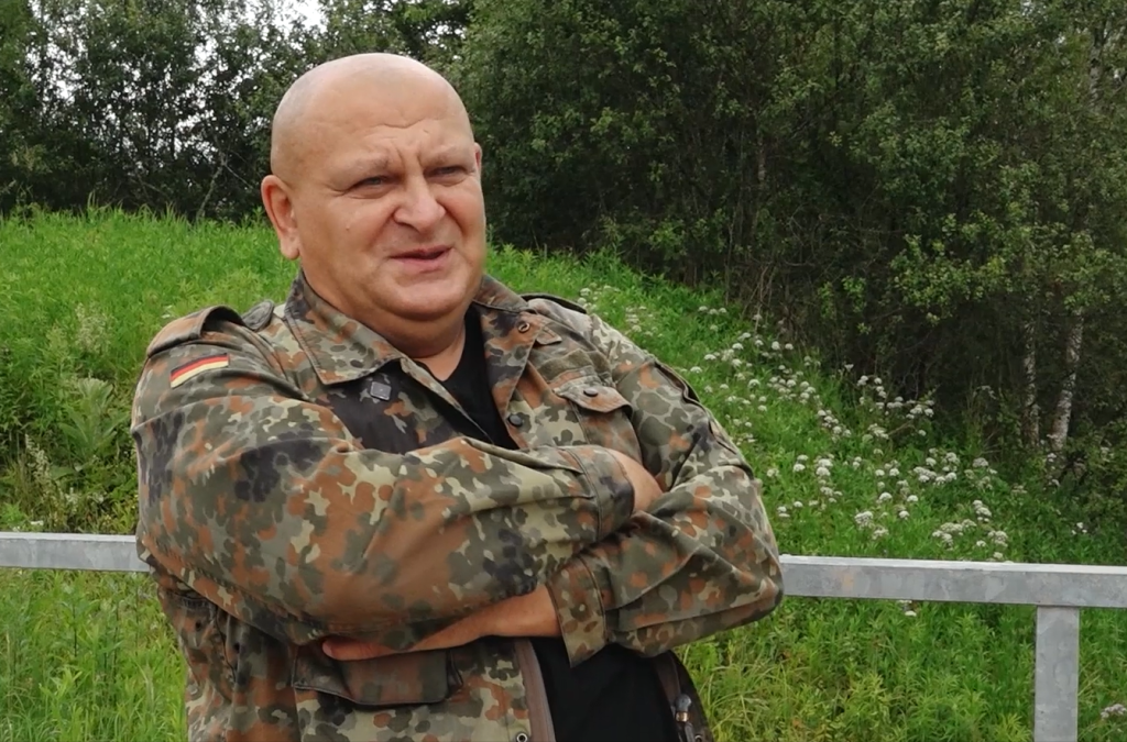 Vīrietis maskēšanās jakā ar sakrustotām rokām Sedas purva dabā