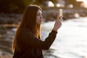 Sieviete ar gariem matiem skatās vai fotografē ar viedtālruni saulrietā vai agrā rīta gaismā.