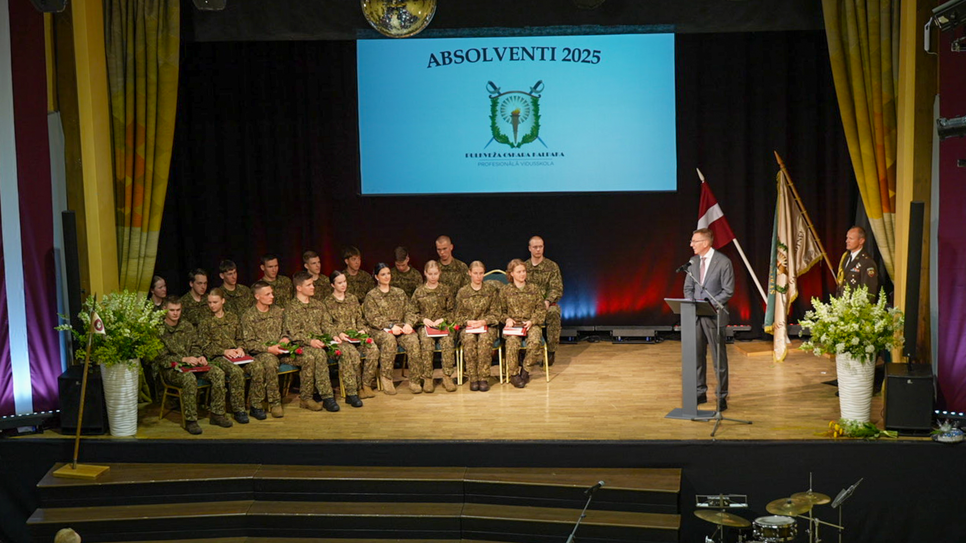 Jaunieši militārās formās sēž uz skatuves, klausoties Valsts prezidenta Edgara Rinkēviča uzrunā, uz ekrāna 