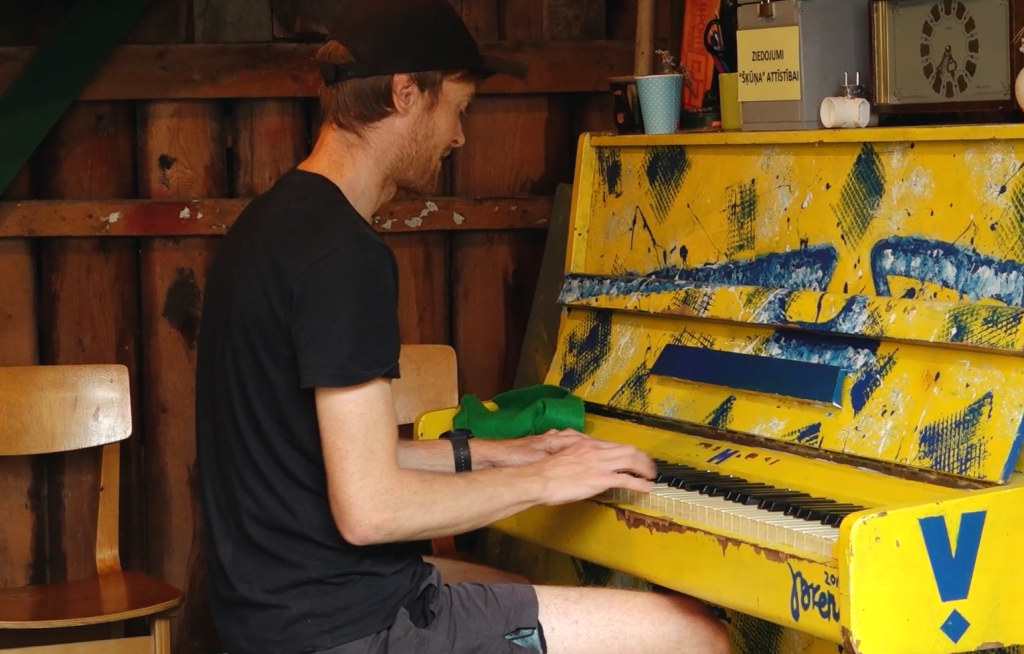 Vīrietis ar melnu cepuri spēlē vecas, dzeltenas klavieres, kas apkrāsotas ar grafiti