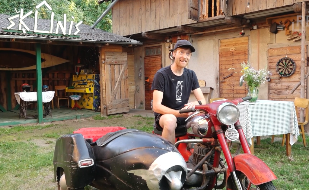 Vīrietis ar smaidu sēž uz veca sarkana motocikla ar blakusvāģi, fonā koka ēkas un uzraksts "ŠĶŪNIS".
