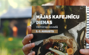 Tuvplāns ar brošūru, uz kuras ir uzraksts "Mājas Kafejnīcu Dienas Valmieras novadā 2.-3. Augusts" un attēloti ēdieni.