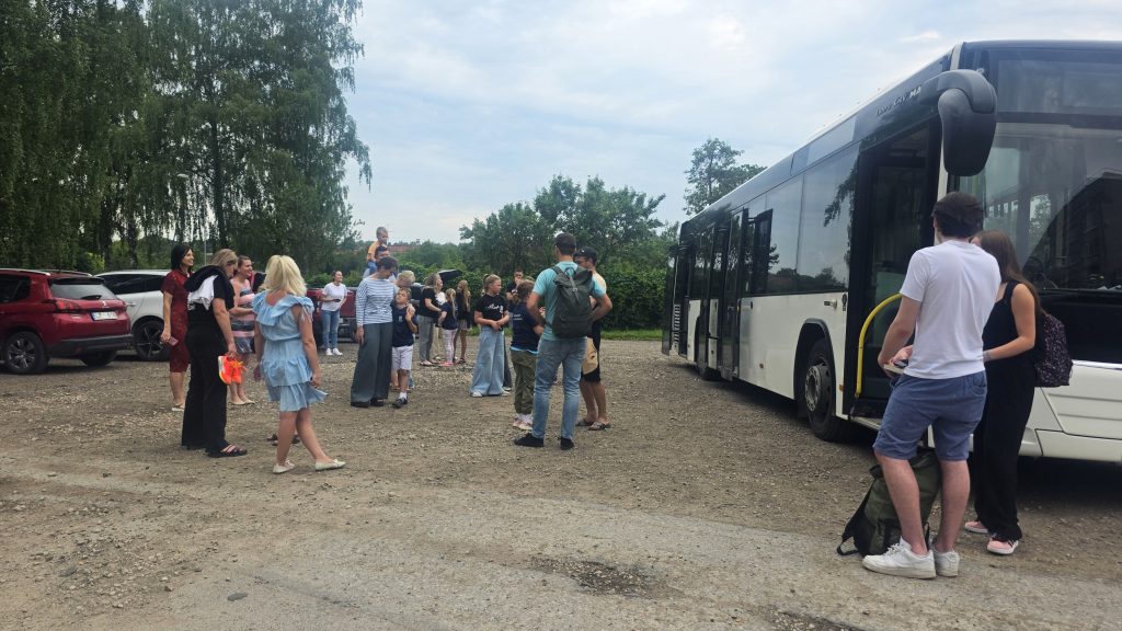 Grupa bērnu un pieaugušo stāv pie liela autobusa pirms brauciena