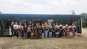 Liela bērnu un pieaugušo grupa stāv rindā pie autobusa un smaida.