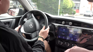 Persona brauc ar automašīnu, turot stūri un skatoties uz priekšu, ar redzamu automašīnas paneli un navigācijas ekrānu.