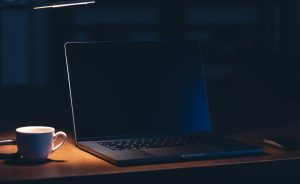 Klēpjdators uz galda blakus kafijas krūzei tumšā telpā ar galda lampu